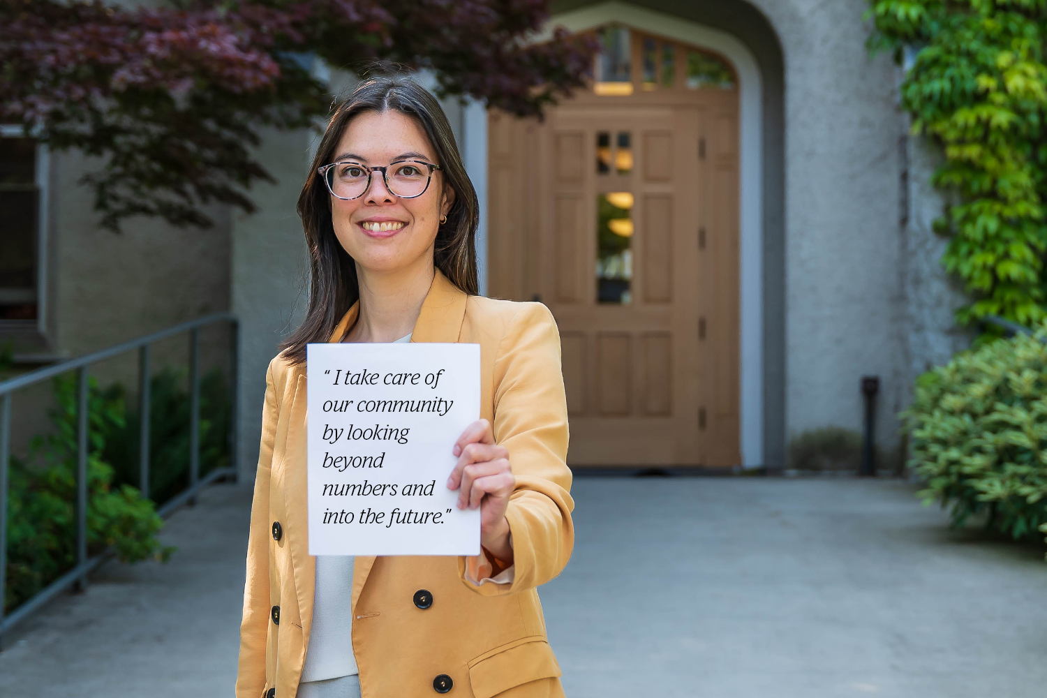 Raffaela stands in front a university building and holds up a white sign that says 'I take care of our community by looking beyond numbers and into the future.'